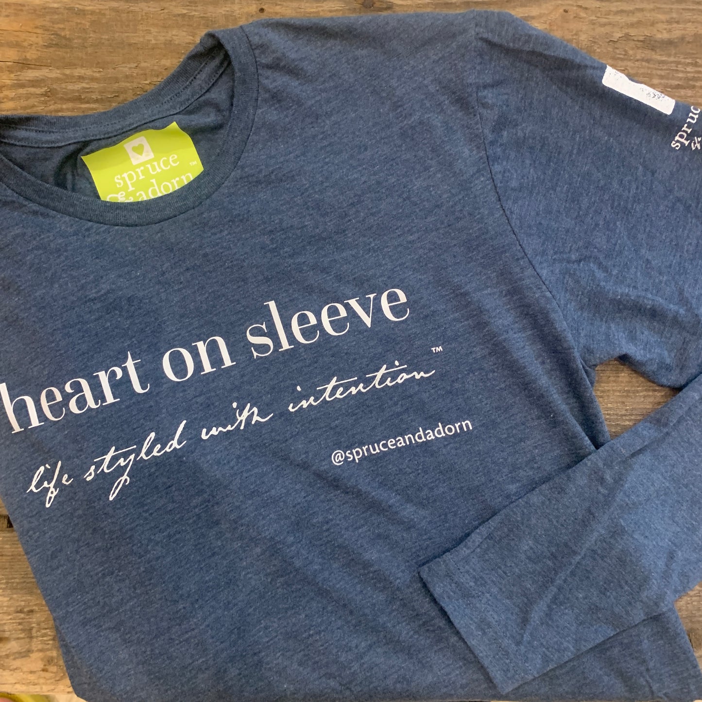 indigo colored signature ‘heart on sleeve’ long sleeve fleece tee reading : ‘heart’ long sleeve tee ,right folded arm with logo and a heart on the right arm patch with signature imprint label laying over a wooden background