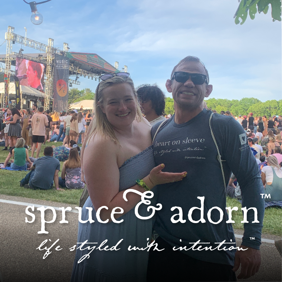 two proud concert goers purchase and wear a 'heart on sleeve' long sleeve tee with arm sleeve tatoo;  in front of center stage; overlayed by spruce & adorn™ life styled with intention™ logo