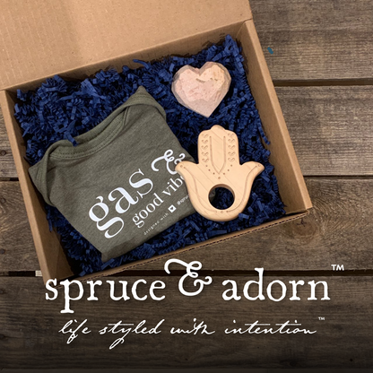 clockwise from left : signature ‘gas & good vibes) baby onsie in color spruce; top right : soothing hand carved soapstone heart; lower right : organic wood hamsa teether in a cardboard box with blue crinkle paper over a wooden background