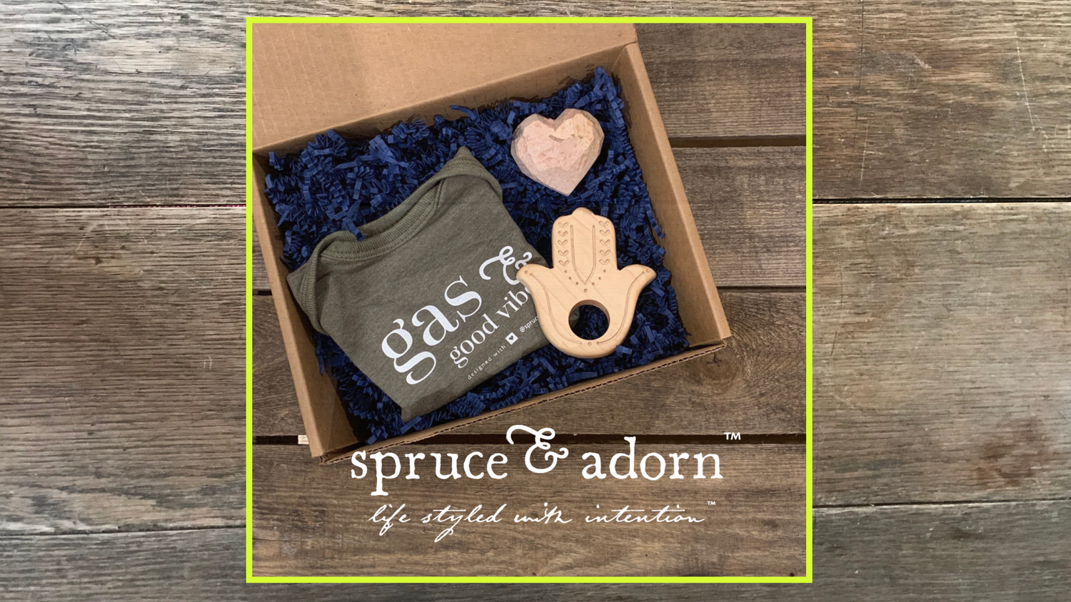 clockwise from left : signature ‘gas & good vibes) baby onsie in color spruce; top right : soothing hand carved soapstone heart; lower right : organic wood hamsa teether in a cardboard box with blue crinkle paper over a wooden background